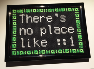 A finished and framed cross stitch that says "There's no place like ::1" with 1s and 0s acting as a decorative border
